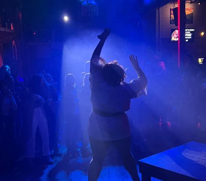 Silhouette of a dancer striking a high-energy pose on a smoke-filled, blue-lit nightclub dance floor under a disco ball, surrounded by a watching crowd and colorful stage lights.