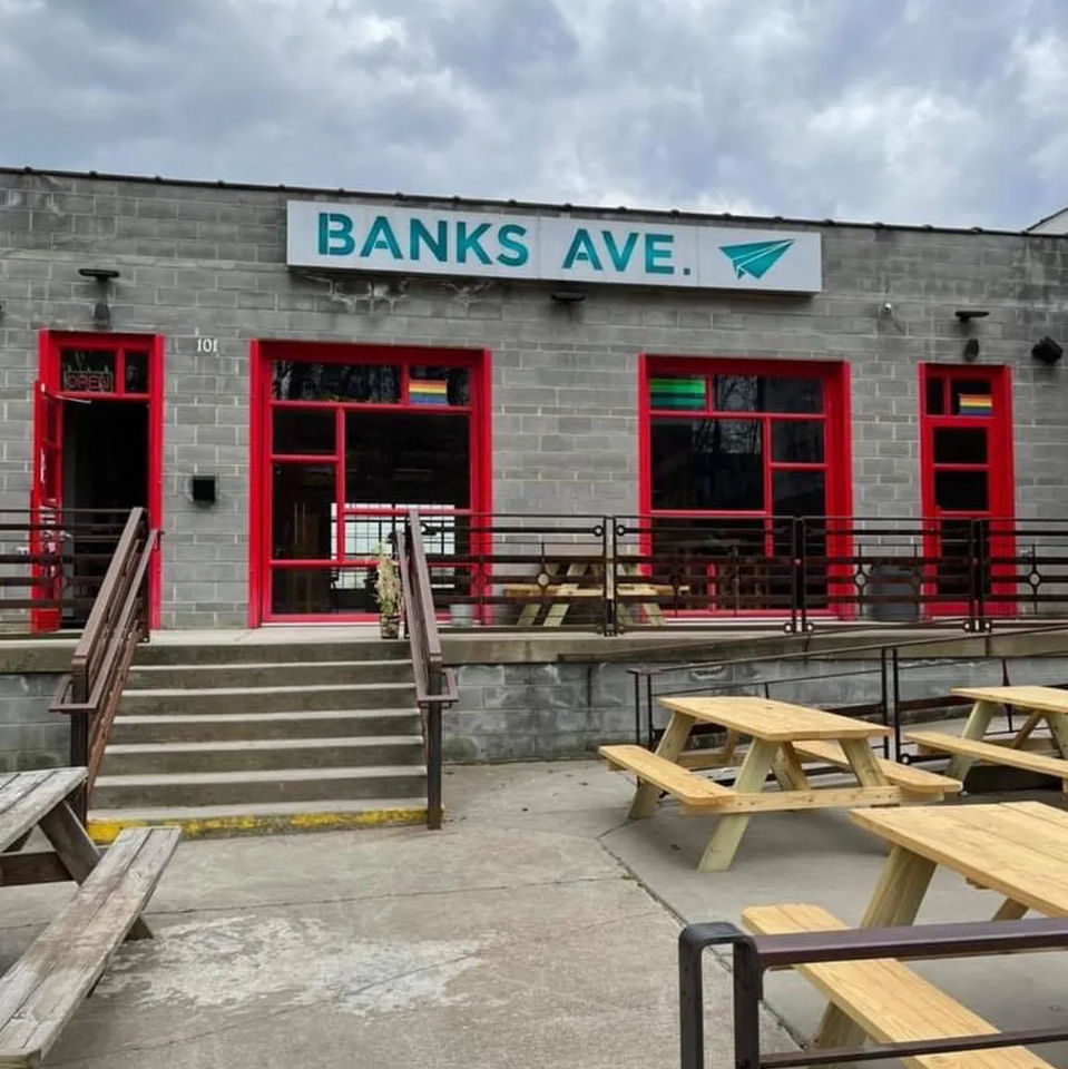 Gray cinderblock storefront with teal sign, red-framed windows and concrete steps leading to an outdoor patio with wooden picnic tables under a cloudy sky.