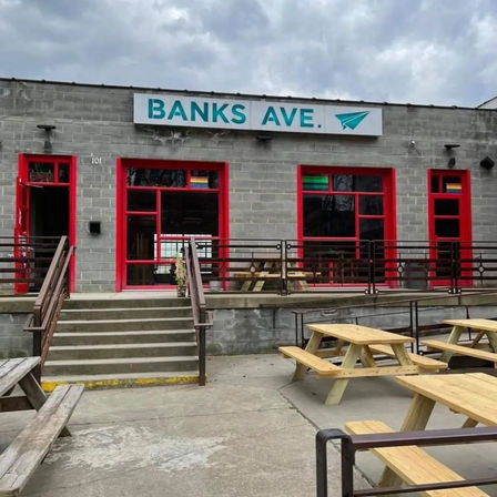Gray cinderblock storefront with teal sign, red-framed windows and concrete steps leading to an outdoor patio with wooden picnic tables under a cloudy sky.