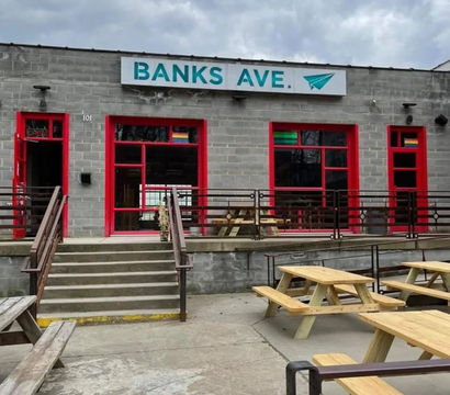 Gray cinderblock storefront with teal sign, red-framed windows and concrete steps leading to an outdoor patio with wooden picnic tables under a cloudy sky.