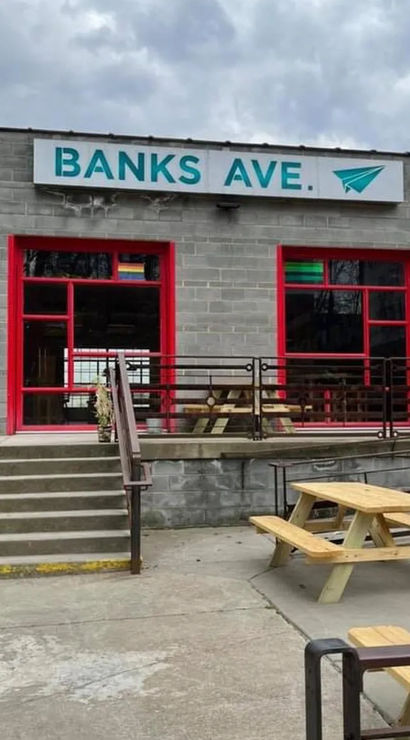 Gray cinderblock storefront with teal sign, red-framed windows and concrete steps leading to an outdoor patio with wooden picnic tables under a cloudy sky.