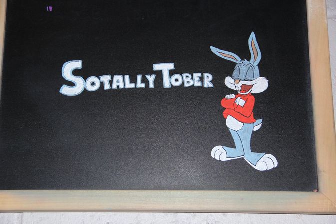 Hand-painted chalkboard sign reading "SOTALLY TOBER" with a grinning cartoon rabbit in a red sweater at right and a light wood frame