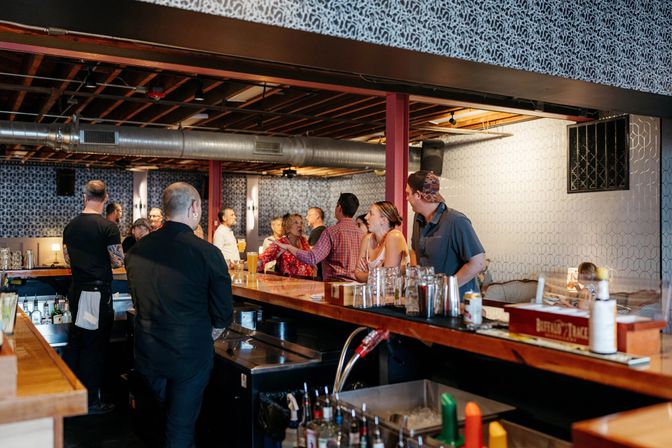 Busy urban bar interior with patrons chatting at a long wooden counter, bartenders serving drinks beneath exposed ceiling ductwork and geometric patterned wallpaper — lively evening scene.