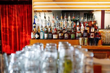 Vibrant cocktail bar interior with rows of liquor bottles on a wooden backbar, mirrored shelves, blurred glassware in the foreground, and a red velvet curtain backdrop.