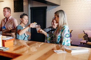 Group of adults toasting cocktails at a polished wooden bar in a cozy lounge with geometric wallpaper