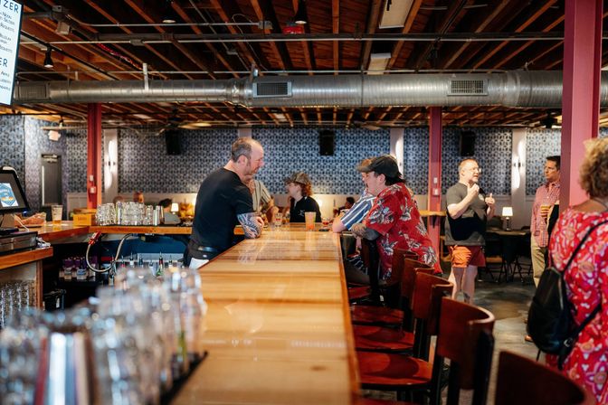 Urban taproom interior with a long polished wooden bar, bartender chatting with patrons on stools, glasses and beers on the counter, exposed ductwork and warm ambient lighting.