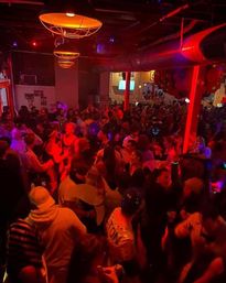 Packed nightclub dance floor under warm industrial pendant lights, red and purple club lighting, a dense crowd dancing and socializing at a lively late-night party with balloons overhead.