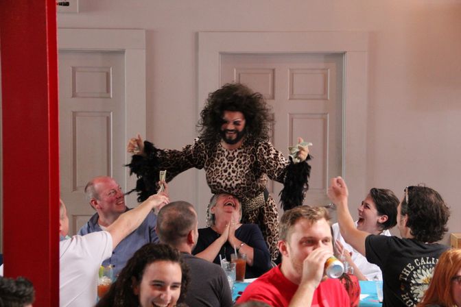 Energetic drag performer in leopard-print dress and big curly hair collecting tips from a laughing indoor brunch crowd, audience members cheer and hand over dollar bills.