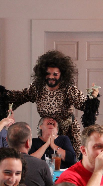 Energetic drag performer in leopard-print dress and big curly hair collecting tips from a laughing indoor brunch crowd, audience members cheer and hand over dollar bills.