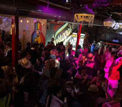 Crowded indoor bar and dance floor with people partying under disco balls and chandeliers, colorful neon mural and DJ booth lit in pink and purple lights.
