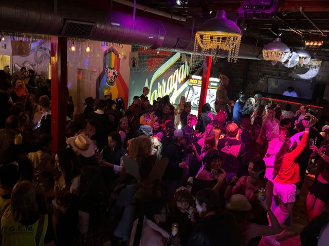 Crowded indoor bar and dance floor with people partying under disco balls and chandeliers, colorful neon mural and DJ booth lit in pink and purple lights.