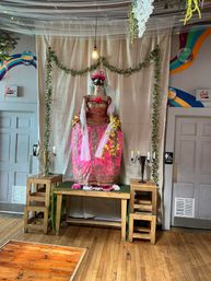 Mannequin wearing an ornate pink and gold ball gown with a black masquerade mask on a decorated indoor photo-op stage, draped fabric backdrop, hanging bulb, ivy garlands, candelabras on wooden stools and hardwood floor