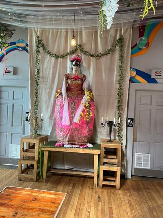 Mannequin wearing an ornate pink and gold ball gown with a black masquerade mask on a decorated indoor photo-op stage, draped fabric backdrop, hanging bulb, ivy garlands, candelabras on wooden stools and hardwood floor