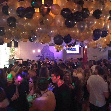 Packed downtown bar party under a ceiling of gold and black balloons, dim purple lighting with a lively crowd dancing and socializing