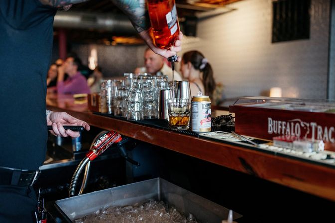 Bartender pouring whiskey into a rocks glass over ice on a wooden bar counter, with stacked glassware, a can of ginger beer and an ice-filled bin in a cozy bar interior.