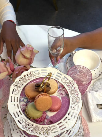 Elegant afternoon tea scene with pastel French macarons on a white ornate two-tiered stand, pink roses, patterned pink glassware, a white teacup, and a manicured hand holding a champagne flute — vintage tea party table setting.