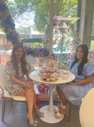 Two women enjoying elegant afternoon tea at a sunlit garden-style patio table with teapot, tiered tray of pastries, and a purple floral garland in the window