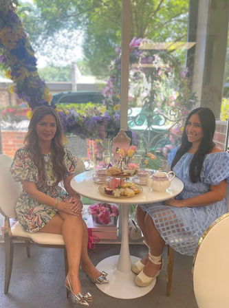 Two women enjoying elegant afternoon tea at a sunlit garden-style patio table with teapot, tiered tray of pastries, and a purple floral garland in the window