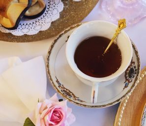 Chic vintage porcelain teacup of black tea on a decorative saucer with an ornate gold spoon, pale pink rose and folded white napkin in an elegant afternoon tea setting