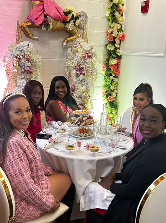 Five women smiling around a round table set for afternoon tea with pastries and champagne, in a pink-and-gold floral party scene with a decorative gold unicorn and flower garlands.