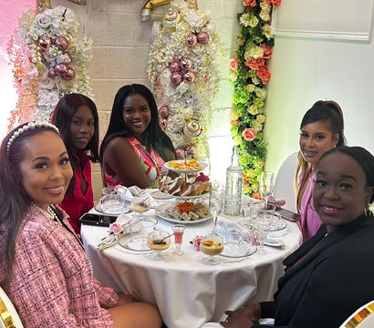 Five women smiling around a round table set for afternoon tea with pastries and champagne, in a pink-and-gold floral party scene with a decorative gold unicorn and flower garlands.