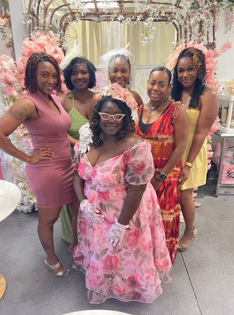 Six women pose at an indoor celebration under a pink blossom arch; the center woman wears a vintage-style pink floral dress, lace gloves, cat-eye glasses and a matching fascinator, surrounded by friends in colorful summer dresses.
