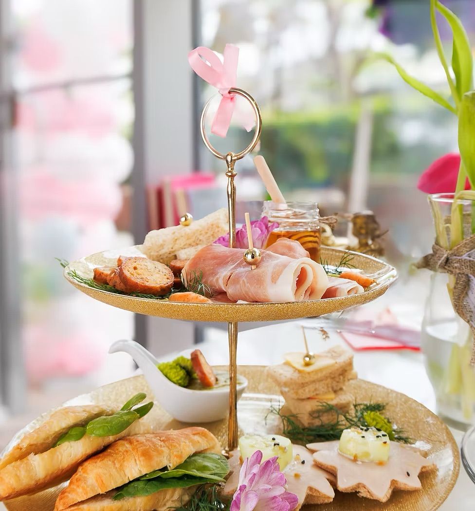 Delightful afternoon tea on a two-tier gold stand with croissant sandwiches, prosciutto, finger sandwiches, honey jar and floral garnish on a sunlit café table