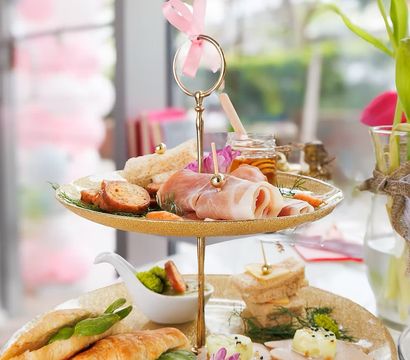 Delightful afternoon tea on a two-tier gold stand with croissant sandwiches, prosciutto, finger sandwiches, honey jar and floral garnish on a sunlit café table