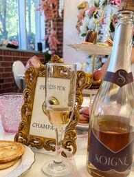 Chic afternoon tea close-up: champagne flute and bottle, ornate gold frame sign reading Press for Champagne, tiered plate of colorful macarons and pink floral decor