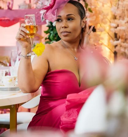 Woman in a fuchsia strapless dress and matching fascinator toasting with a champagne flute garnished with a yellow flower at an elegant indoor floral tea party.