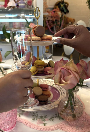 Colorful macarons on a two-tier porcelain stand with a pink rose and hands holding a glass at an elegant afternoon tea setting