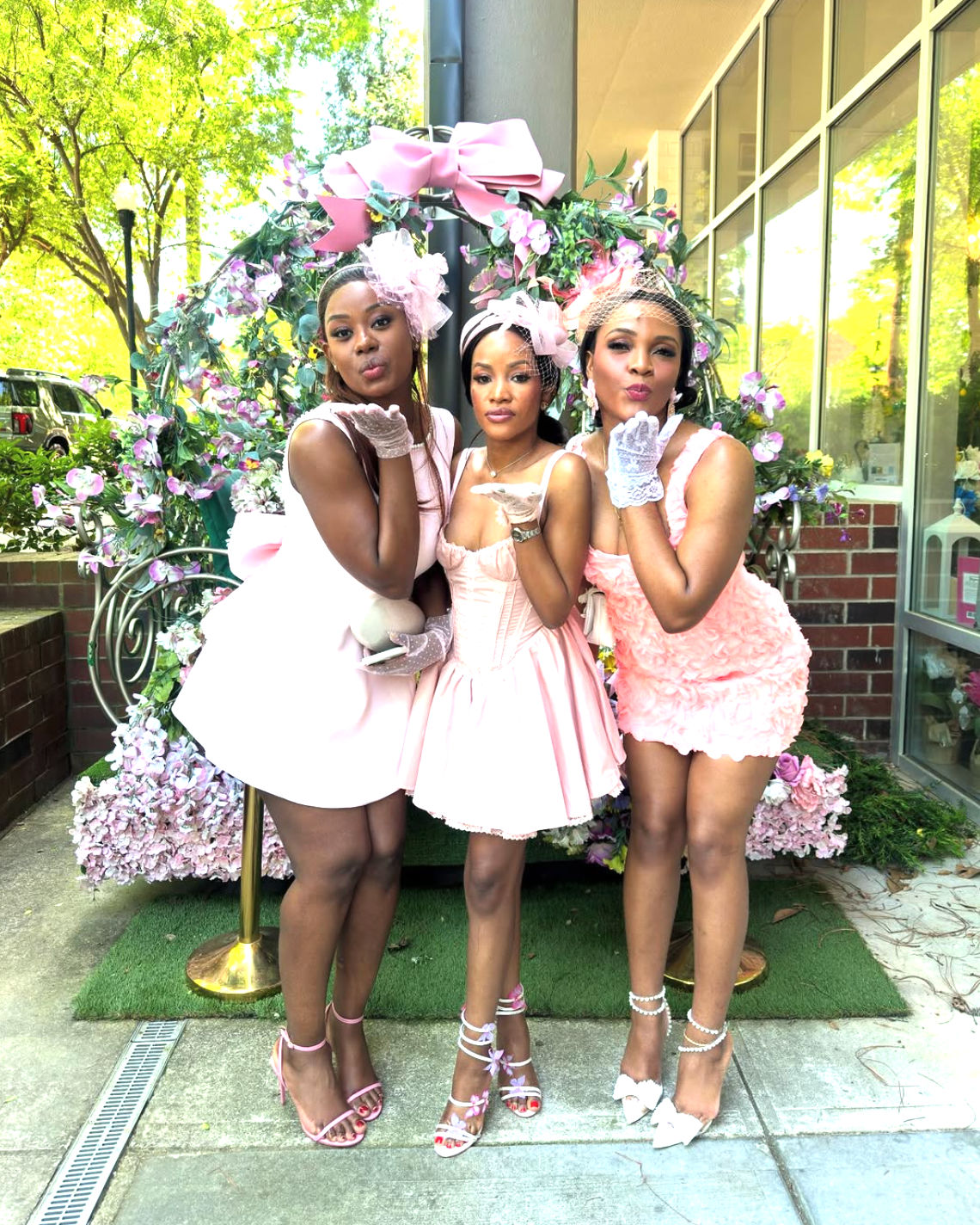 Three women in pink dresses, fascinators and heels playfully blowing kisses in front of a large floral archway outside a storefront on a sunny city sidewalk.