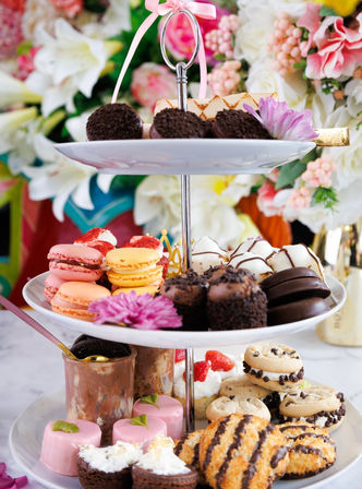 Three-tier dessert stand with colorful macarons, chocolate truffles, mini cakes and assorted cookies on an elegant afternoon tea display against a vibrant floral backdrop