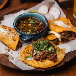 Crispy Mexican-style birria tacos and quesabirria topped with shredded beef, chopped onion and cilantro, served with a blue bowl of savory consommé and radish slices on a rustic wooden platter