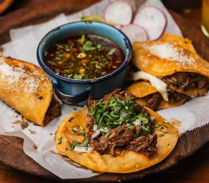 Crispy Mexican-style birria tacos and quesabirria topped with shredded beef, chopped onion and cilantro, served with a blue bowl of savory consommé and radish slices on a rustic wooden platter