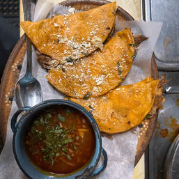 Three golden-orange quesabirria tacos sprinkled with cotija and cilantro on parchment-lined wooden plate, served with a small bowl of savory beef consommé and spoon for dunking