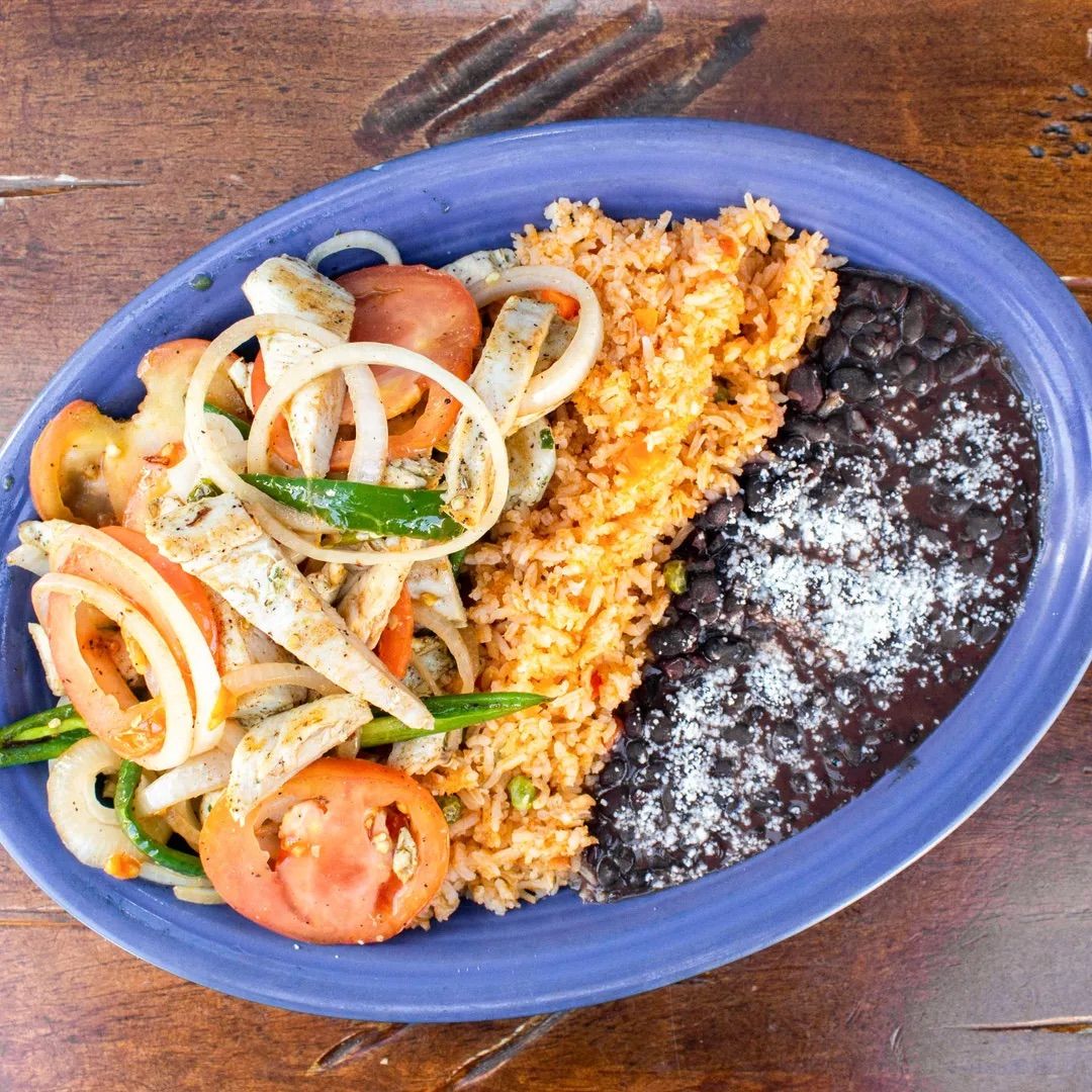 Blue oval plate with colorful grilled chicken fajita strips, sautéed onions, tomatoes and green peppers, Mexican-style rice and black beans topped with crumbled cheese on a wooden table.