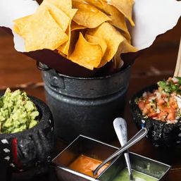 Tortilla chips in a rustic metal bucket with stone bowls of guacamole, pico de gallo and two dipping sauces on a wooden table — Mexican appetizer ready to dip