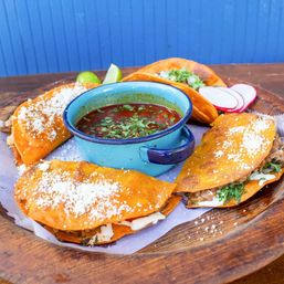 Wooden platter of crispy birria tacos dusted with cotija cheese and cilantro, filled with melted cheese and shredded meat, served with a blue enamel bowl of rich red consommé, lime wedges and sliced radishes against a blue backdrop.