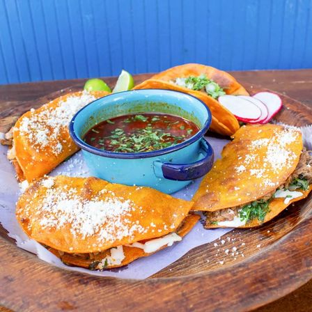 Wooden platter of crispy birria tacos dusted with cotija cheese and cilantro, filled with melted cheese and shredded meat, served with a blue enamel bowl of rich red consommé, lime wedges and sliced radishes against a blue backdrop.