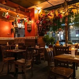 Cozy candlelit Mexican-style restaurant interior with warm orange walls, wooden tables and chairs, decorative tile trim, hanging string lights and small Mexican flags, large floral arrangement and a window overlooking a nighttime city street.