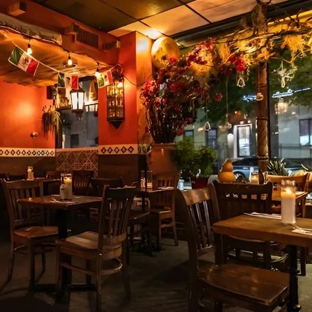 Cozy candlelit Mexican-style restaurant interior with warm orange walls, wooden tables and chairs, decorative tile trim, hanging string lights and small Mexican flags, large floral arrangement and a window overlooking a nighttime city street.