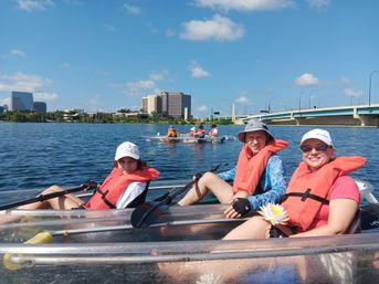 Three people kayaking in clear hull kayaks wearing orange life jackets and sun hats, holding paddles and a white water lily on a sunny urban waterfront with a city skyline and bridge in the background