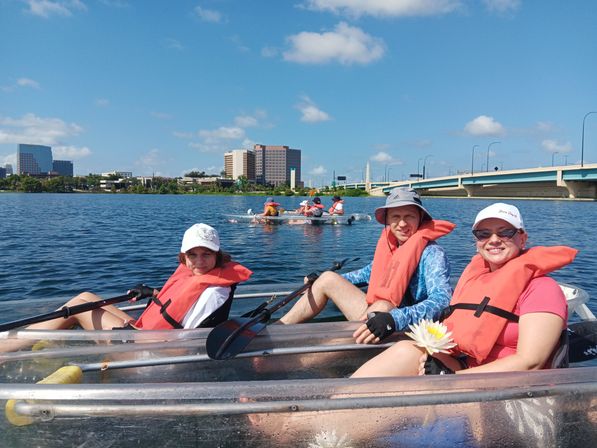 Three people kayaking in clear hull kayaks wearing orange life jackets and sun hats, holding paddles and a white water lily on a sunny urban waterfront with a city skyline and bridge in the background