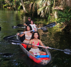 Palm-lined tropical creek with a group kayaking — two women paddling a clear red tandem kayak ahead of two paddlers, lush green foliage surrounding the water.