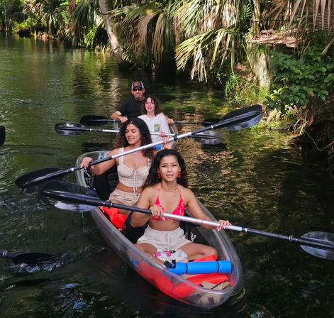 Palm-lined tropical creek with a group kayaking — two women paddling a clear red tandem kayak ahead of two paddlers, lush green foliage surrounding the water.