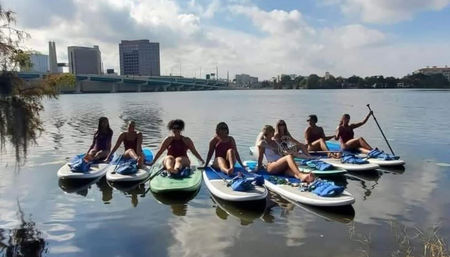 Group of eight paddlers lounging on stand-up paddleboards in a calm urban river, life jackets on the boards, with a bridge and downtown skyline reflected on the water under a partly cloudy sky.