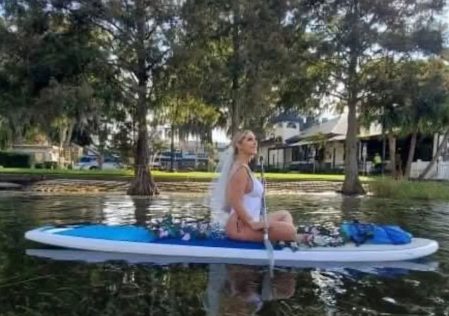 Bride in white dress and veil sitting cross-legged on a flower-adorned paddleboard on a calm lake with waterfront homes and trees in the background.