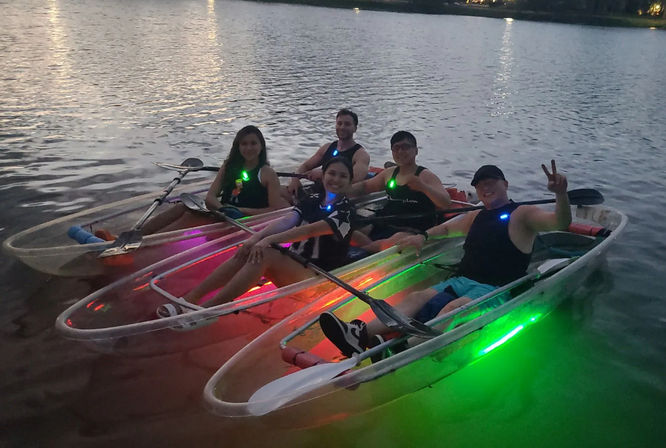 Five people in transparent kayaks on a calm lake at dusk, colorful LED lights (green, red, purple) glowing beneath the boats as paddles rest across laps and one person flashes a peace sign — evening group kayaking fun.