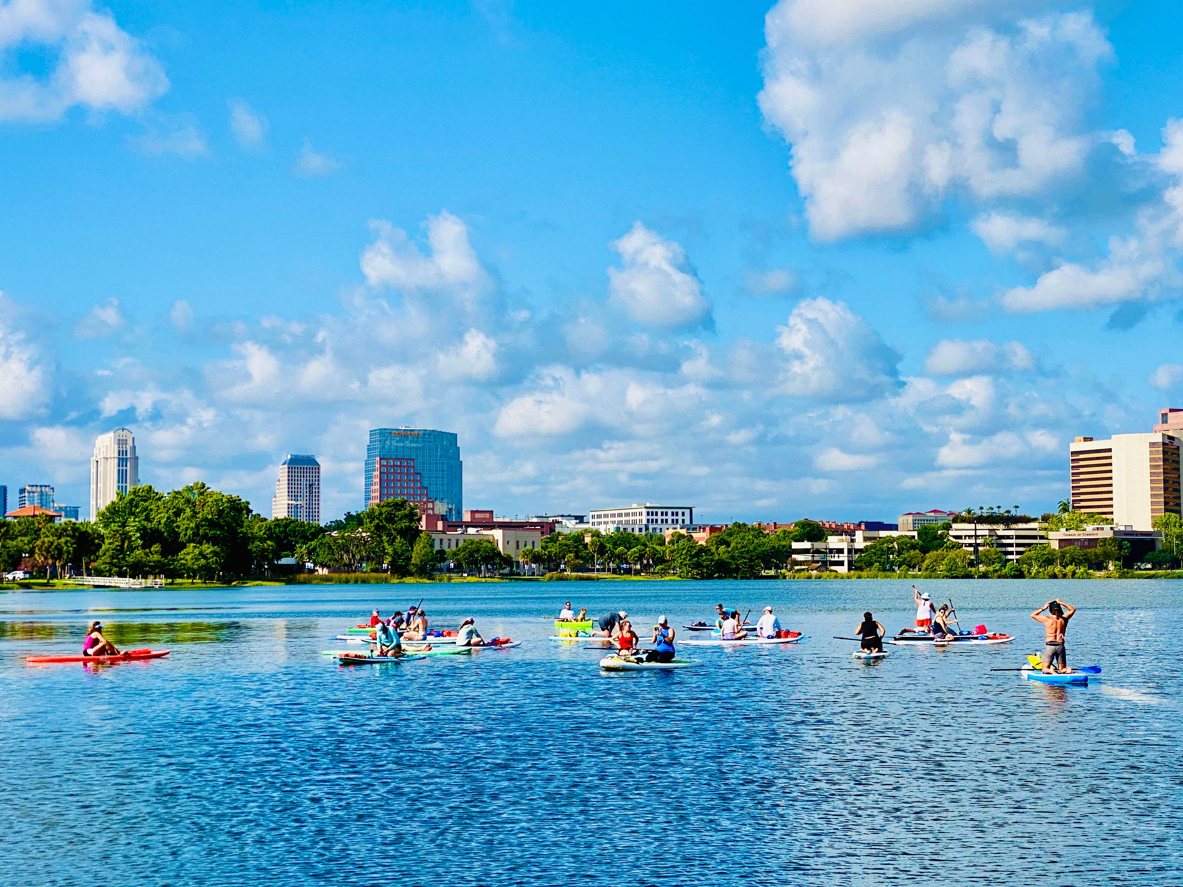 Paddleboarders and kayakers enjoying a sunny urban lake with downtown skyline, green shoreline, and blue sky dotted with puffy clouds